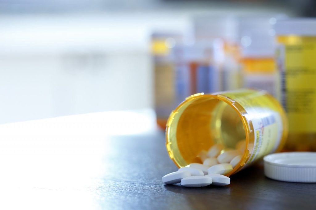 Pills pour out of a prescription medication bottle that lays on its side onto a kitchen counter as a strong morning light filters in through a window. Several other pill bottles stand out of focus in the background.
