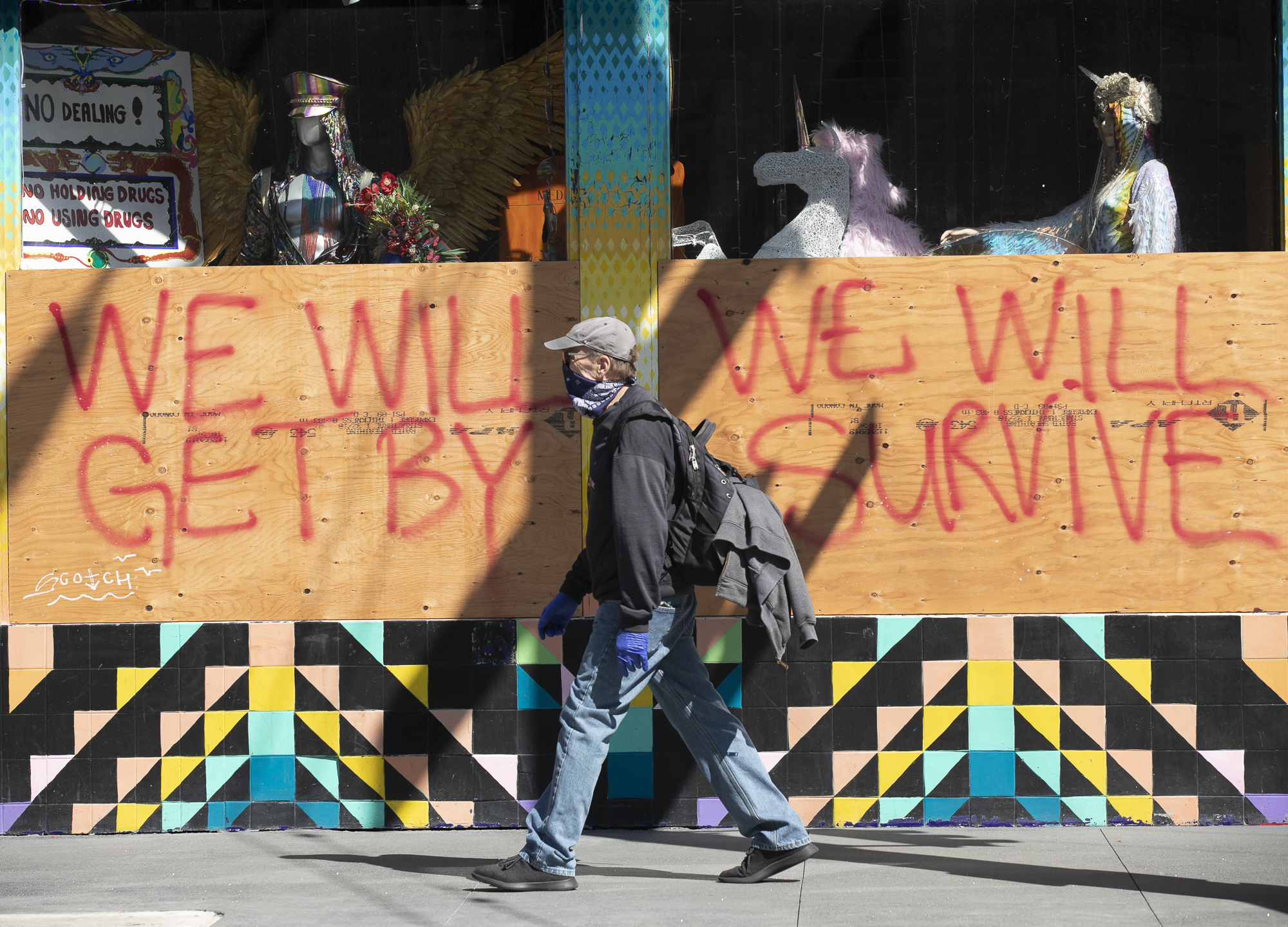 A man wearing disposable gloves and a bandana over his face walks past a boarded up shop on Haight Street in San Francisco on April 7, 2020. California is entering the fourth week of sheltering in place to limit the spread of the novel coronavirus, and the number of Californians hospitalized and those in ICUs with COVID-19 is starting to edge up only slightly. Photo by Anne Wernikoff for CalMatters