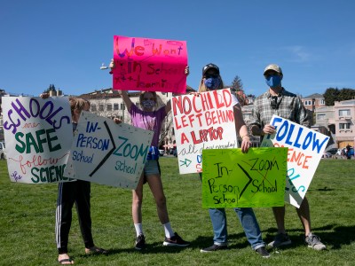 From left: Harrison, 8, and Allie, 10 carry signs with their parents Elizabeth Owen and Steve Ganotis during a rally to reopen Oakland public school for in-person instruction at Astro Park on Feb. 28, 2021. Photo by Anne Wernikoff, CalMatters