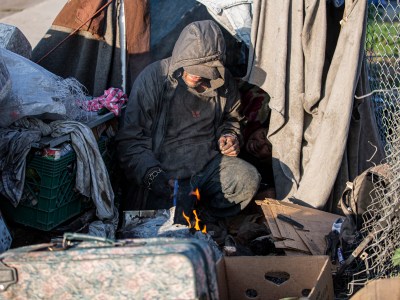 A man starts a fire in his makeshift tent along a barbed wire fence near Highway 99 in southwest Fresno on Feb. 11, 2022. The fence blocks out a grass area that used to be a homeless encampment. Photo by Larry Valenzuela for CalMatters.