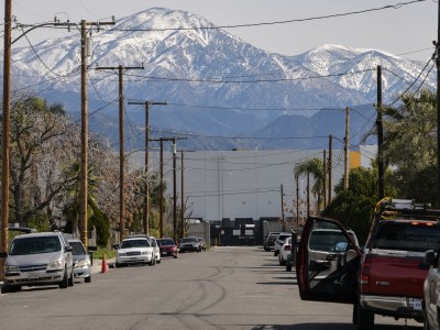A large warehouse sits in front of snow capped mountains in a residential neighborhood. A low perspective shows a line of cars parked a long the street.