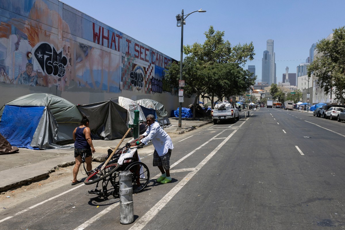 A homeless man wearing a white button up shirt and plaid shorts pushes a wheelchair next to a homeless encampment in Skid Row with the Los Angeles skyline in the background.