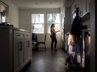 Soft daylight enters the kitchen through windows as an adult person stands near a fridge, while in the foreground a child with long hair, a purple dress, and roller-skates leans on a countertop.