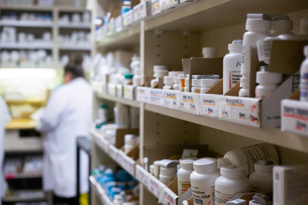 Prescription drugs in bottles and boxes on shelves at a pharmacy, while pharmacists work in the background.