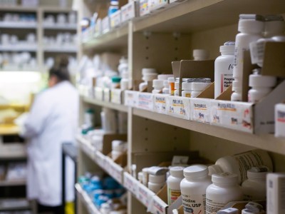 Prescription drugs in bottles and boxes on shelves at a pharmacy, while pharmacists work in the background.