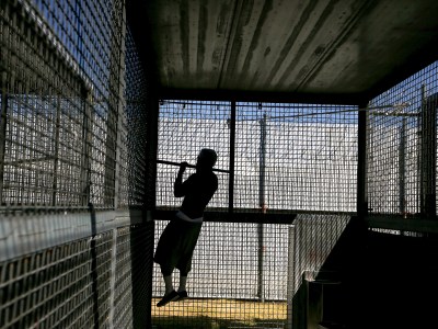 A shirtless inmate, with shorts and tennis shoes, does a pull-up on a bar inside of an outdoor-cage in a prison yard in Sacramento. Fencing and razor wire is surrounds the entire cage.