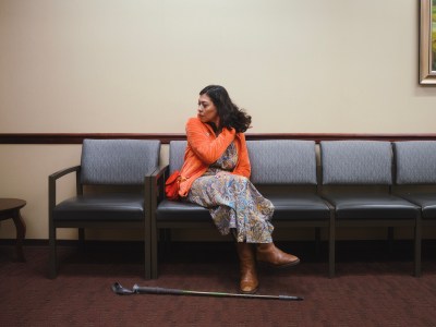 A person in a bright orange fleece sweater and a paisley dress sits on a chair inside a waiting room at a doctors office in Pasadena. A walking cane lays on the floor in front of them.