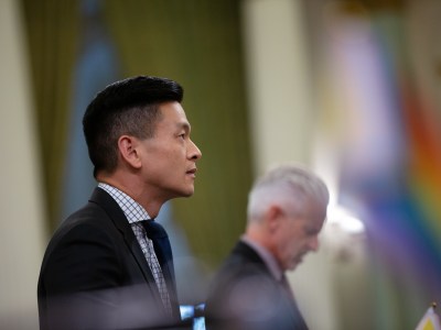 The profile of Assemblymember Evan Low with another lawmaker in the distance and a out-of-focus rainbow flag on the edge of the frame.