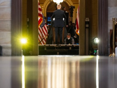 A rearview of a person standing at a podium. They are framed by a door and inside the room cameras and press face the person. American flags are visible at the side, inside the room. The photo is from a ground-level perspective. Lights reflect off the polished ground. The setting is a press conference.