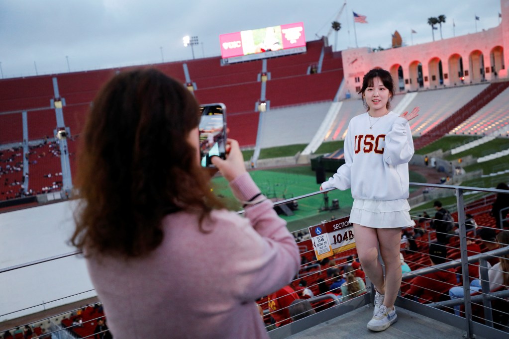A person on the bottom left side of the frame holds up their phone to take a photo of another person wearing a white sweatshirt with the embroidered letters "USC" at a commencement ceremony at the Los Angeles Memorial Coliseum in Los Angeles.