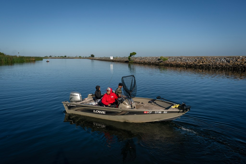 Three people sit in a fishing boat while in the Sacramento-San Joaquin Delta. The rivers' bank is visible on the edge of the photo. The light is soft, evoking dawn or sunset.