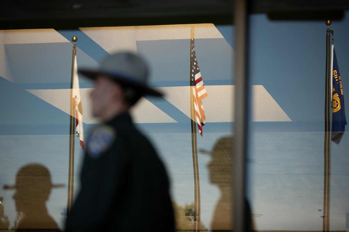 The reflection of a law enforcement officer is seen on a window along with three flag poles with the U.S., California and CHP flags.