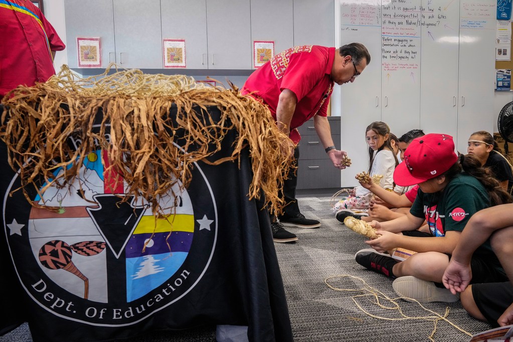 Assemblymember James C. Ramos, in a red shirt, leads a cultural activity with a group of children sitting on the floor, working with woven materials. A table covered with straw and a 'Dept. of Education' banner is visible in the foreground.