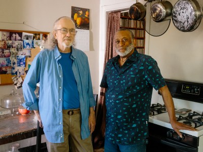 Luke Johnson and husband Osbey Jackson stand in the kitchen of their home. Jackson rests his hand on a kitchen stove, and Johnson leans back on a chair next to a kitchen table. The home is decorated with photos and pots and pans hang over the stove.