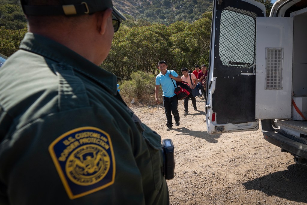 A U.S. Customs and Border Protection officer, identifiable by a patch on their uniform, stands near an open van door, observing a group of people approaching. The scene is outdoors in a rural area with dry, rugged terrain and sparse vegetation. The individuals walking towards the van carry personal belongings and appear to be guided or escorted by the officer.