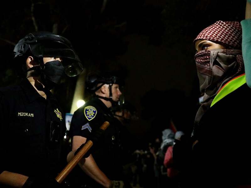 A police officer wearing riot gear and holding a baton stands inches away from a protester wearing a red and white patterned keffiyeh and a safety vest during a protest.