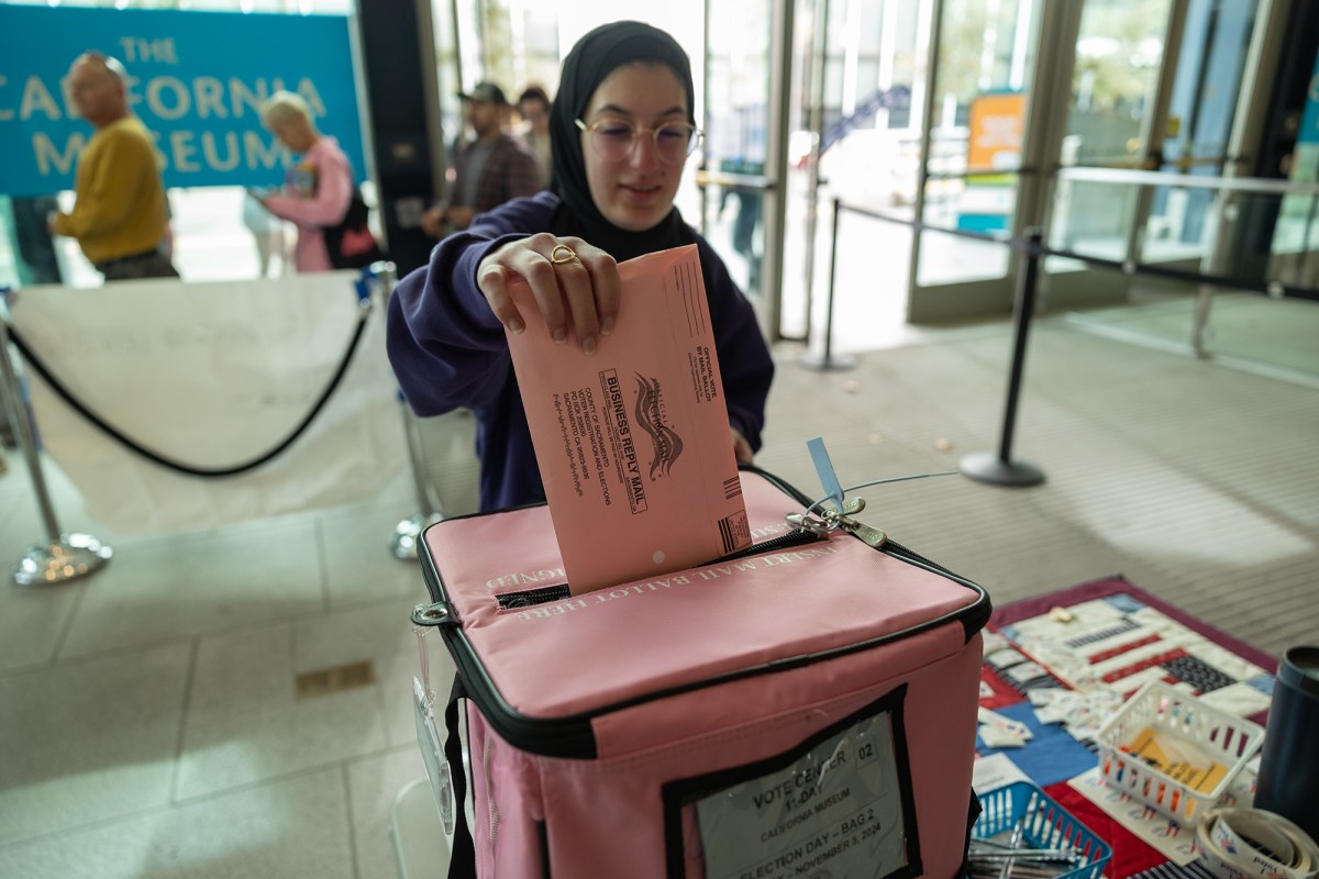 A person wearing a hijab and purple sweatshirt inserts a pink ballot into a pink ballot box at a polling place.