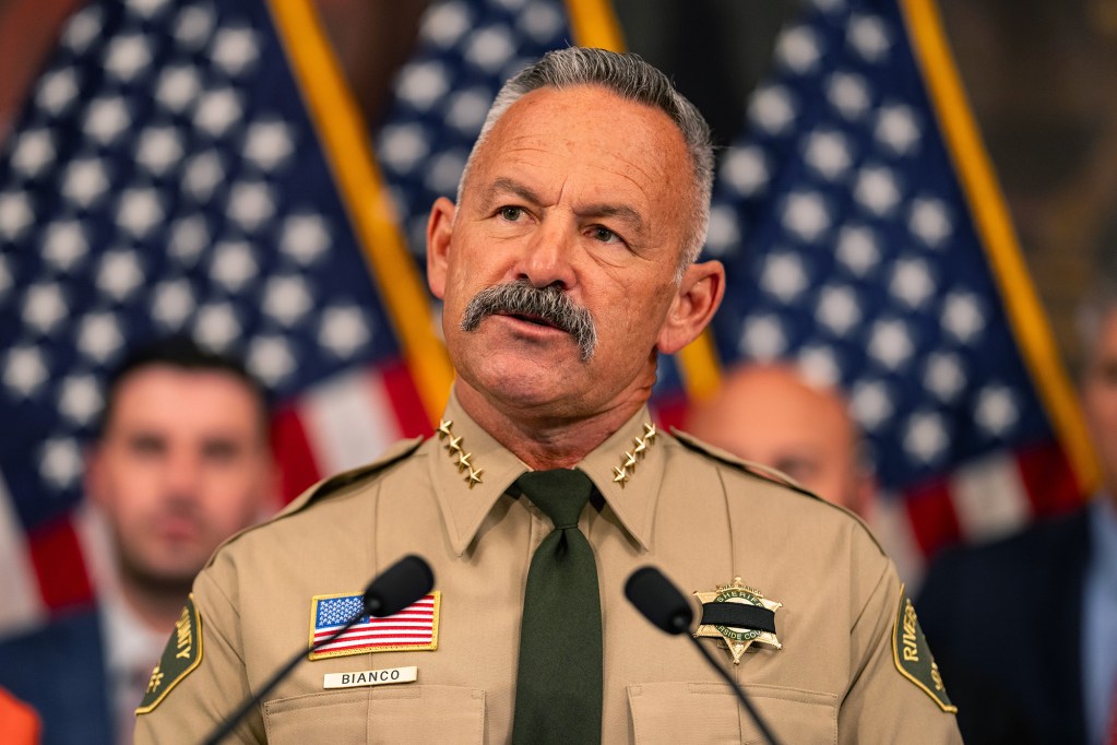 A close-up view of a person wearing a brown sheriff's uniform standing behind a lectern with two small microphones. Three people can be seen standing behind him next to three American flags.