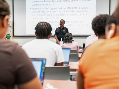 Students look toward the front of a classroom while a professor stands and lectures.