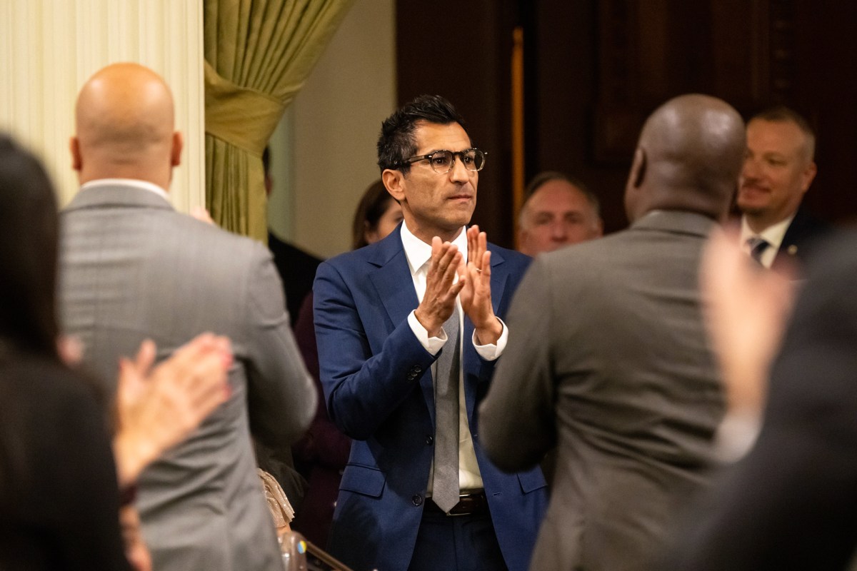 A person wearing a blue suit stands up while clapping as other people face him and clap.
