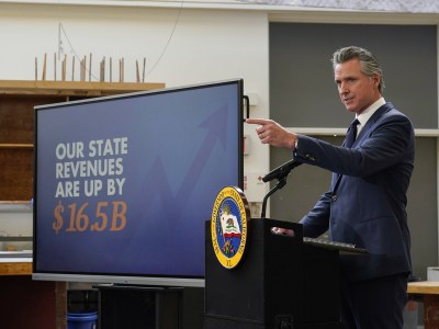 A person wearing a navy blue suit points in front of them as they stand behind a lectern with the California seal in front of it. A large screen with a graphic that reads "our state revenues are up by $16.5 B" is seen next to him.