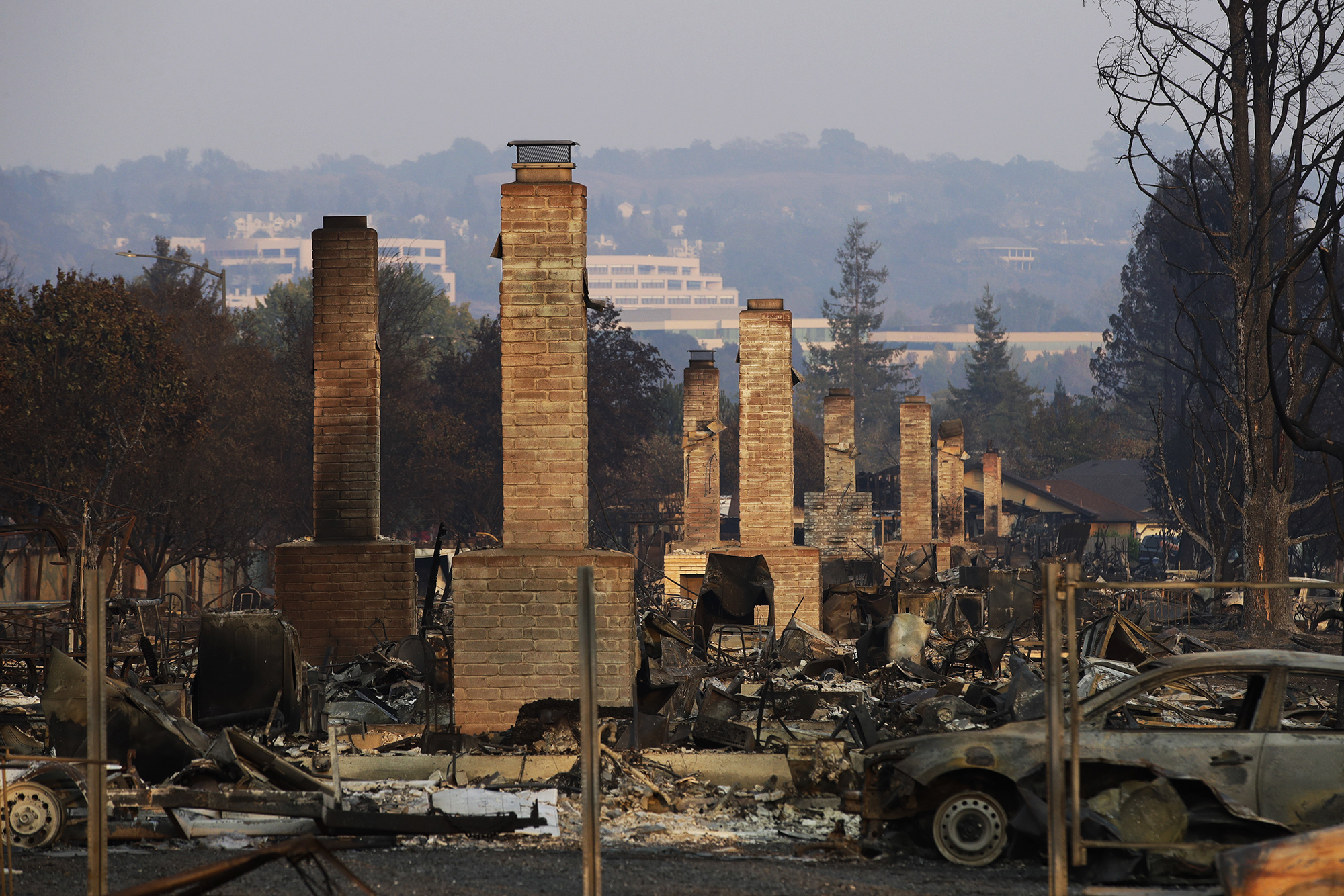 A row of eight chimneys remain standing amongst the debris of a neighborhood destroyed by the Tubbs Fire. The city of Santa Rosa is in the background, along with burned-down trees, in the foreground, debris and a burned vehicle on the street.