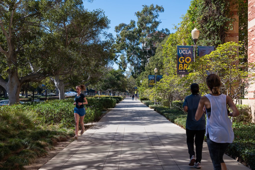 Three people run on a tree-lined sidewalk at a college campus on a sunny day. UCLA banners can be seen on lamp posts the right side of the sidewalk.