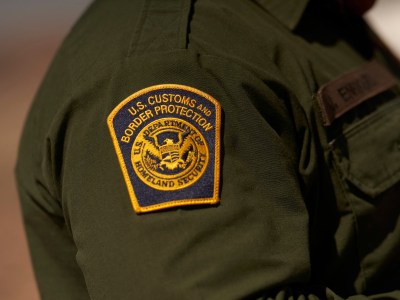 A close-up view of a U.S. Customs and Border Protection patch on the left arm of an agent's green uniform. The patch contains the words "U.S. Customs and Border Protection" and "U.S. Department of Homeland Security" and its logo, which includes an eagle's breast as a shield divided into three sections containing elements that represent homeland - air, land, and sea.