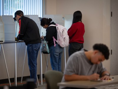 Individuals are seen voting at private booths in a well-lit room, with one person seated at a nearby table, filling out paperwork. Some voters have backpacks and are focused on the process. The background includes vertical window blinds and a neutral interior setting.