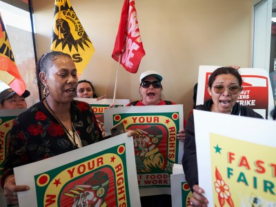A group of fast food workers protest inside a restaurant, holding signs and flags advocating for workers' rights. Some individuals are chanting, while others display expressions of determination. The signs feature bold colors and text, including phrases like 'Your Rights' and 'Fast Fair.' A yellow flag with the words 'Fast Food Workers' is visible in the background.