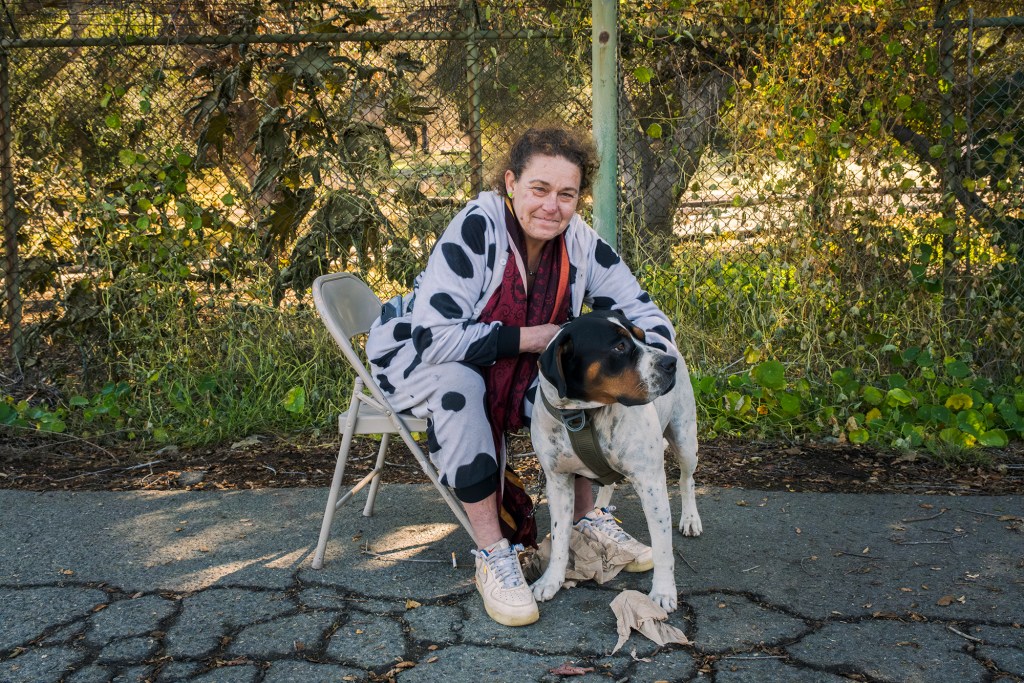 A person sits on a folding chair on a cracked asphalt surface, smiling while holding a large dog with a harness. They are wearing a polka-dotted onesie and sneakers, with a red scarf draped around their neck. The dog stands attentively, looking off to the side. Behind them, a chain-link fence covered in overgrown greenery stretches across the background.