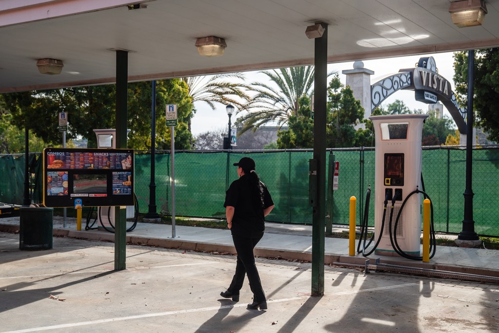 The back of a person wearing a black hat, shirt and pants as they walk by a whitel electric vehicle charging station at a drive-thru Sonic location.