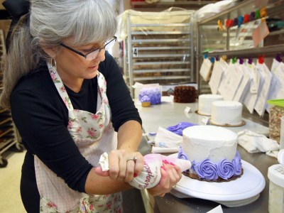 A baker in a floral apron decorates a cake with purple frosting roses using a piping bag in a commercial kitchen. Several cakes in various stages of decoration are visible on the stainless steel work surface, along with clipped order sheets, baking tools, and storage containers. Metal racks filled with trays are in the background.
