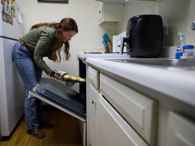 A person wearing a green long-sleeve blouse, a brown buckle belt and blue jeans leans forward as they insert a cupcake tray into the oven of their small kitchen. The person can barely bend over because the refrigerator is so close to the oven.