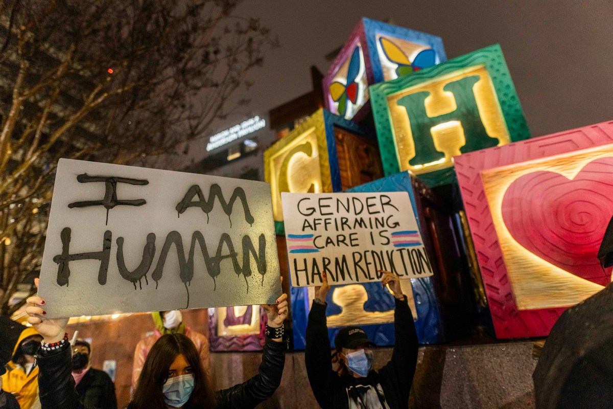 Two people wearing blue surgical masks hold signs in front of a giant letter block sculpture outside a hospital during a recent evening. One sign reads "I am Human" and the other reads "Gender Affirming Care is Hard Reduction" and includes two drawings of the transgender flag.