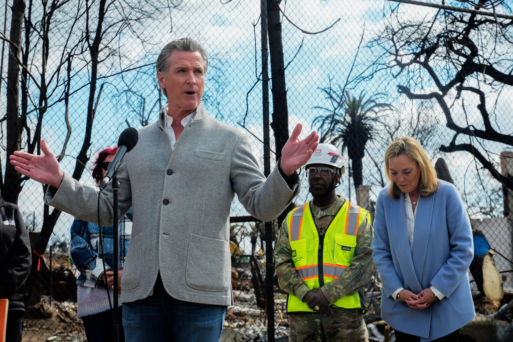 A person wearing a grey jacket stands behind a microphone in front of a group of people behind them. In the background is a chained linked fence with burned trees and homes behind it.