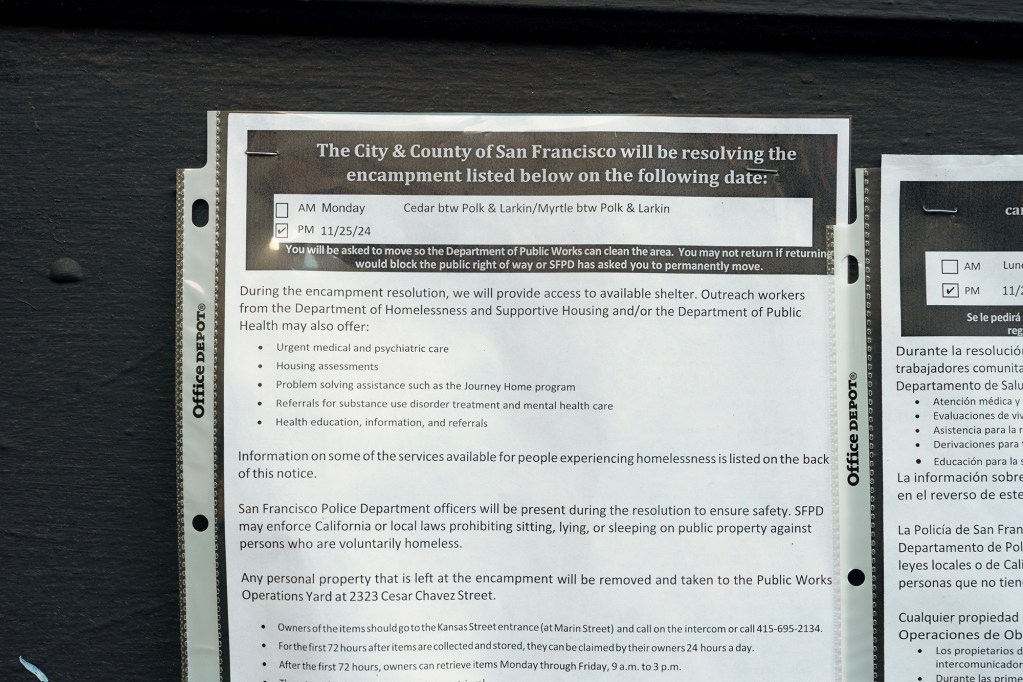 A notice from the City and County of San Francisco is pinned to a black surface, informing residents about the resolution of a homeless encampment. The document, placed in a clear plastic sleeve, details the date and location of the clearance, services available for displaced individuals, and the involvement of the police department. The paper is bordered by additional notices in English and Spanish, providing similar information.