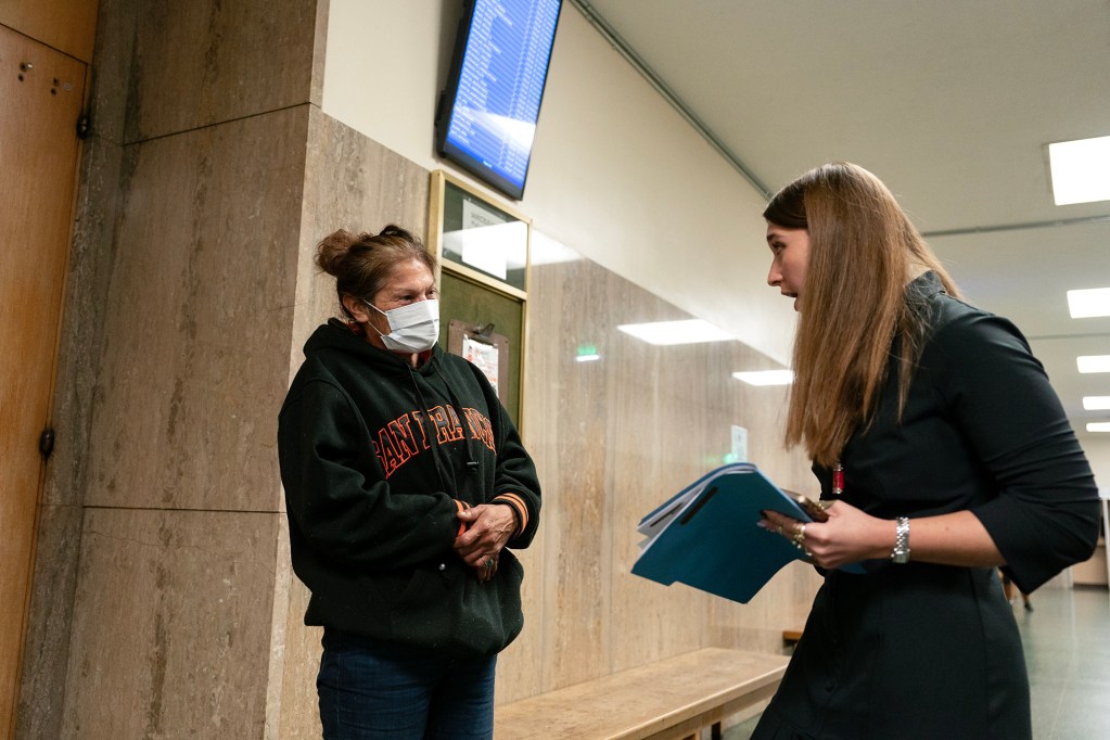 A woman in a black professional outfit holds a blue folder and speaks with a woman in a black hoodie with "San Francisco" written across the front in orange letters. The woman in the hoodie wears a face mask and stands with her hands clasped together. They are in a courthouse hallway with beige marble walls, a wooden bench, and an electronic court docket screen overhead displaying case information. The conversation appears serious and focused.