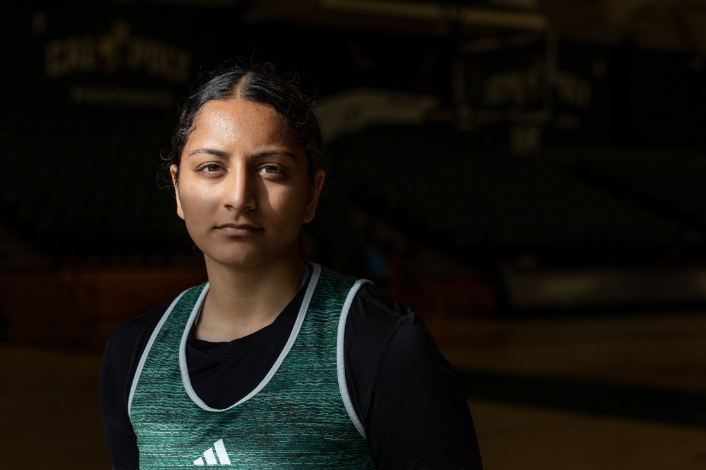 A basketball player wearing a green Cal Poly jersey stands in a dimly lit gymnasium, with dramatic lighting highlighting their face. They have a focused expression, looking directly at the camera. The background is dark, with faintly visible bleachers and the Cal Poly Mustangs logo, emphasizing the subject in the foreground.