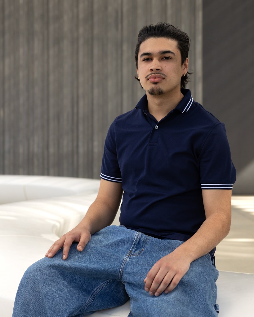 A portrait of a person as they sit on a white concrete bench against a gray backdrop. The person has slick-back black hair and wears a dark blue collar shirt and light blue jeans.
