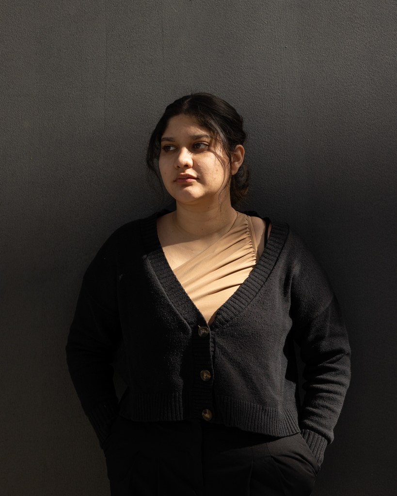 A portrait of a person standing against a black backdrop and soft rays of light shine on their right cheek and upper body. The person has brown curly hair tied in a bun and wears a black cardigan with a champagne-colored blouse.