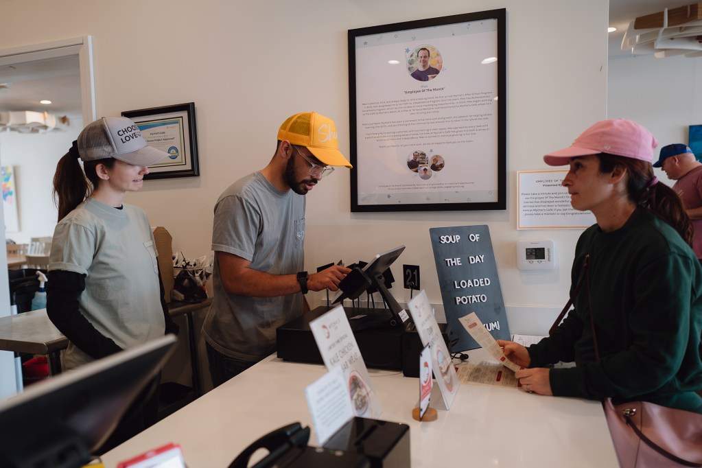 A left-side view of a worker, wearing a yellow hat and gray shirt as another worker stands next to them wjile they help take a customers order.
