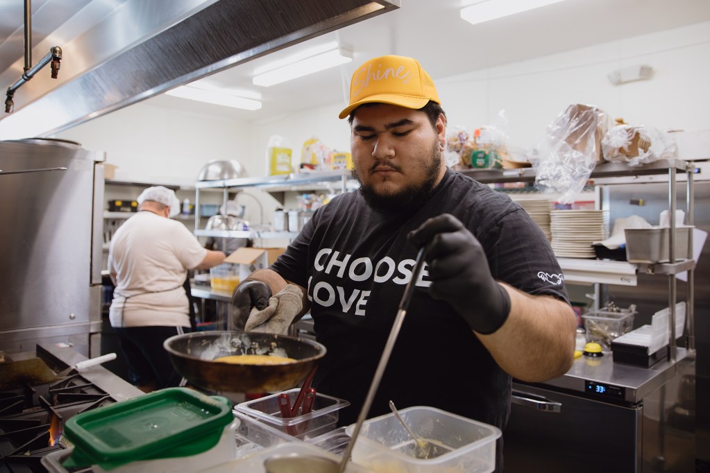 A person, wearing a yellow hat and black shirt with the words "Choose Love" on it, gets ready to pour a yellow liquid into a saucepan in the kitchen of a cafe.
