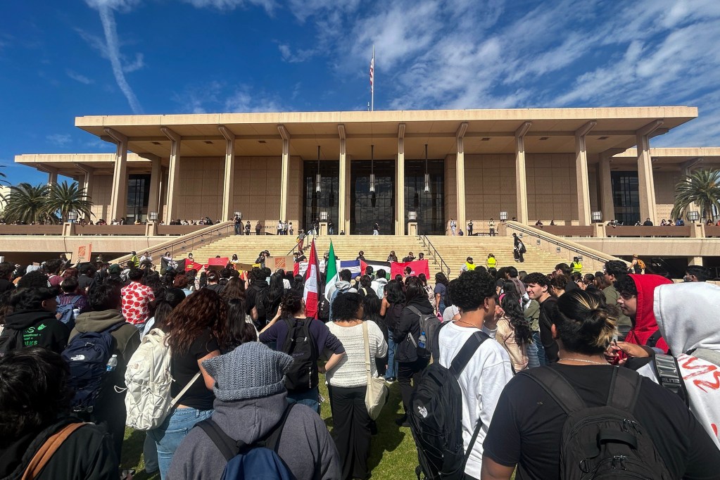 A large crowd gathers on a lawn in front of a grand, modern building with tall columns and a wide staircase. Many individuals carry backpacks, and some wear hooded sweatshirts or head coverings. Several people hold up red signs and flags, including a green, white, and red flag. Security personnel in yellow vests stand along the steps, while others observe from the upper level. The sky is bright blue with wispy clouds, and palm trees line the sides of the scene.