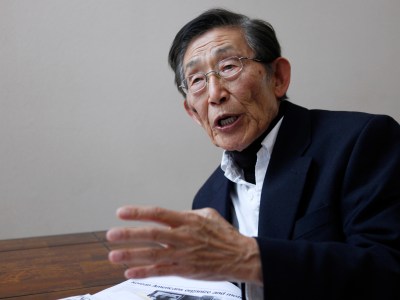 An elderly person, wearing gray reading glasses, a black suit jacket with a white collar shirt, gestures with their right hand as they speak next to a table with print newspaper copies.