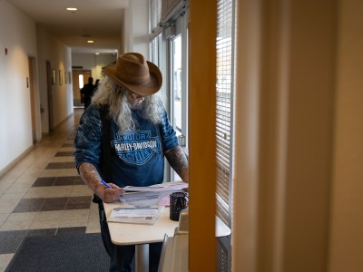 A person wearing a brown cowboy hat, glasses, and a Harley-Davidson t-shirt stands at a small table inside a hallway, filling out paperwork with a pen. Their long, wavy hair and tattoos are visible as they focus on the documents. The setting is a public building with tiled floors, beige walls, and a row of doors leading to other rooms. Natural light streams in through the windows, which have partially closed blinds. A few people are visible in the background walking through the hallway.