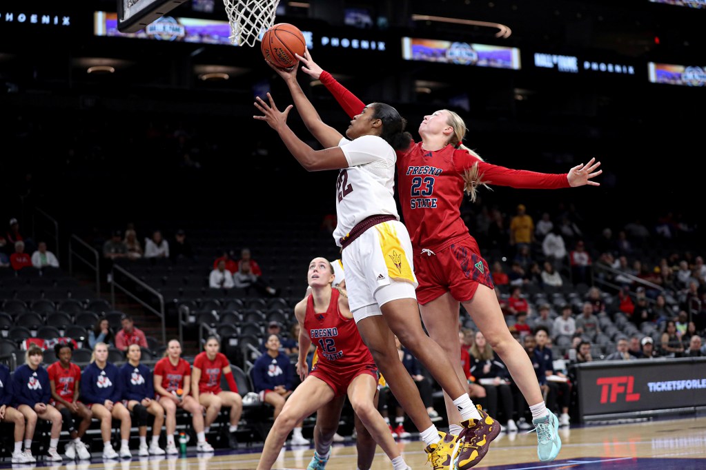 A basketball player in a white and gold uniform leaps for a layup while a defender in a red Fresno State jersey attempts to block the shot. The player in white extends their arm toward the hoop, gripping the basketball, while the defender stretches out with an outstretched arm. Other players and team members sit on the bench in the background, watching the play. The game takes place in an indoor arena with spectators visible in the stands.
