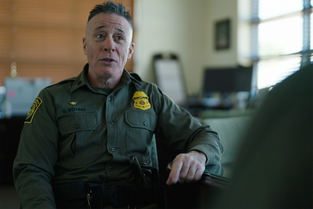 A uniformed U.S. Border Patrol agent sits in an office during an interview, speaking and gesturing with one hand. The nameplate on the uniform reads "G. Bovino." The background shows office furniture and a window with blinds partially open.