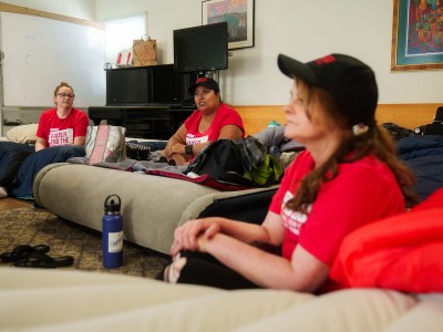 Three women wearing matching red shirts sit on air mattresses in a room as they participate in a hunger strike.
