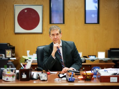 A person in a suit sits at a wooden desk in an office, looking directly at the camera with one hand resting near their chin. The desk is cluttered with office supplies, personal photos, and decorations. Behind them, a framed artwork featuring a large red circle hangs on a wood-paneled wall, flanked by tall vertical windows.
